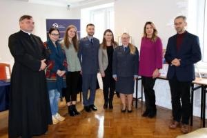 Na zdjęciu widać kilkoro stojących osób, w tym funkcjonariuszy Służby Więziennej i kapelana więziennego.
